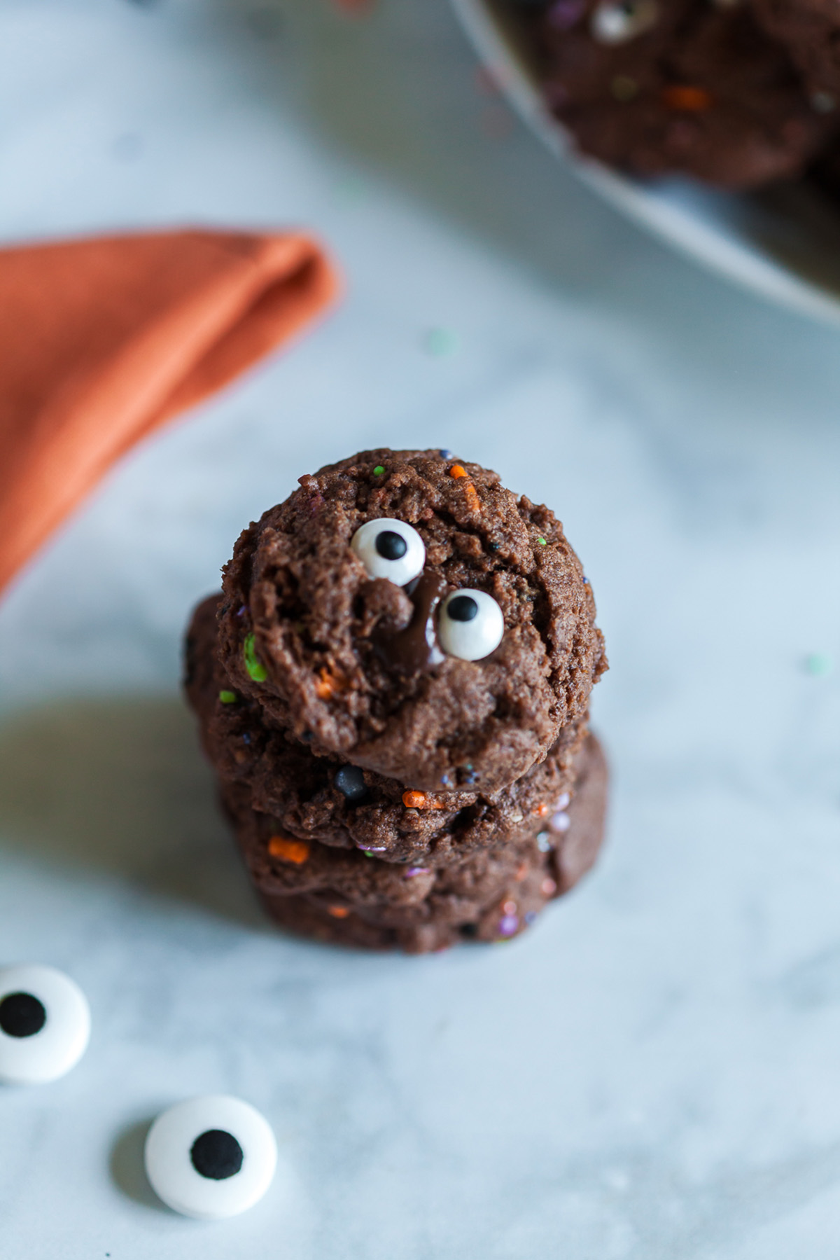 Three stacked chocolate Halloween cookies decorated with candy eyes, with more candy eyes scattered nearby.