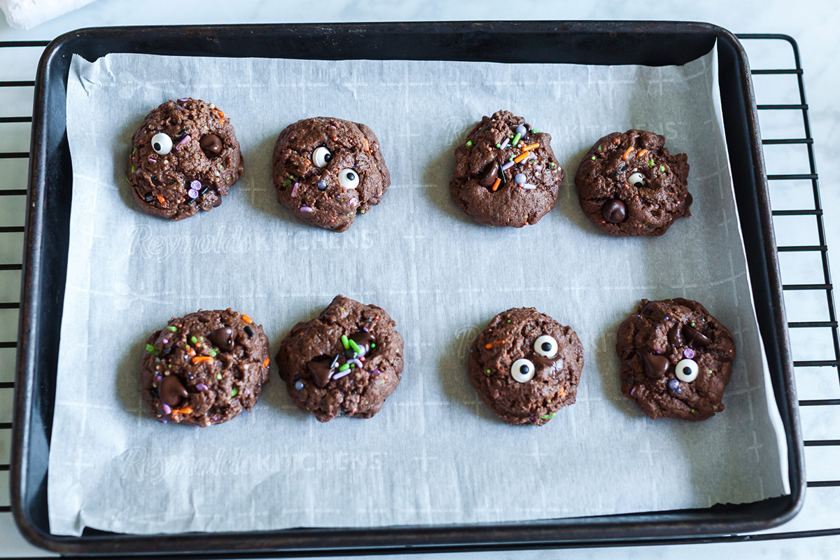 Baked chocolate Halloween cookies on a parchment-lined baking sheet topped with sprinkles and candy eyes.