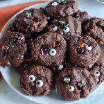 Close-up of chocolate Halloween cookies topped with candy eyes, sprinkles, and melted chocolate chips on a white plate.