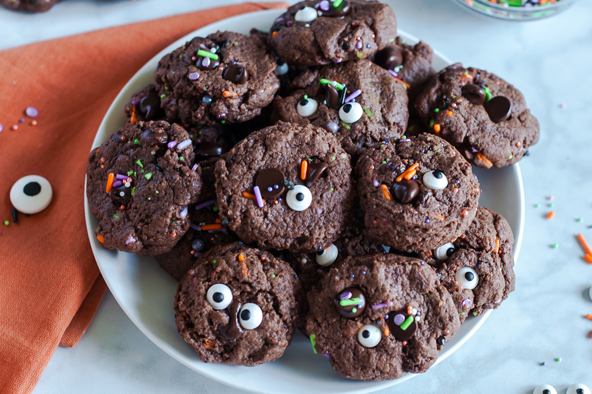 Close-up of chocolate Halloween cookies topped with candy eyes, sprinkles, and melted chocolate chips on a white plate.