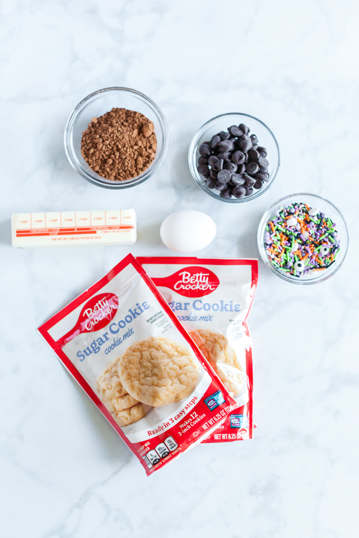 Ingredients for chocolate Halloween cookies including sugar cookie mix, cocoa powder, butter, egg, chocolate chips, and Halloween sprinkles.