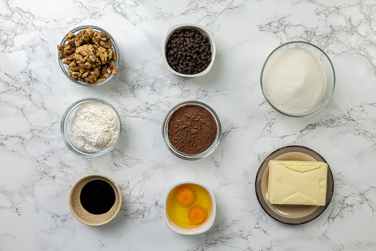Bowls filled with ingredients for chocolate walnut brownies including cocoa powder, flour, walnuts, sugar, butter, eggs, chocolate chips, vanilla and baking powder on a marble surface.