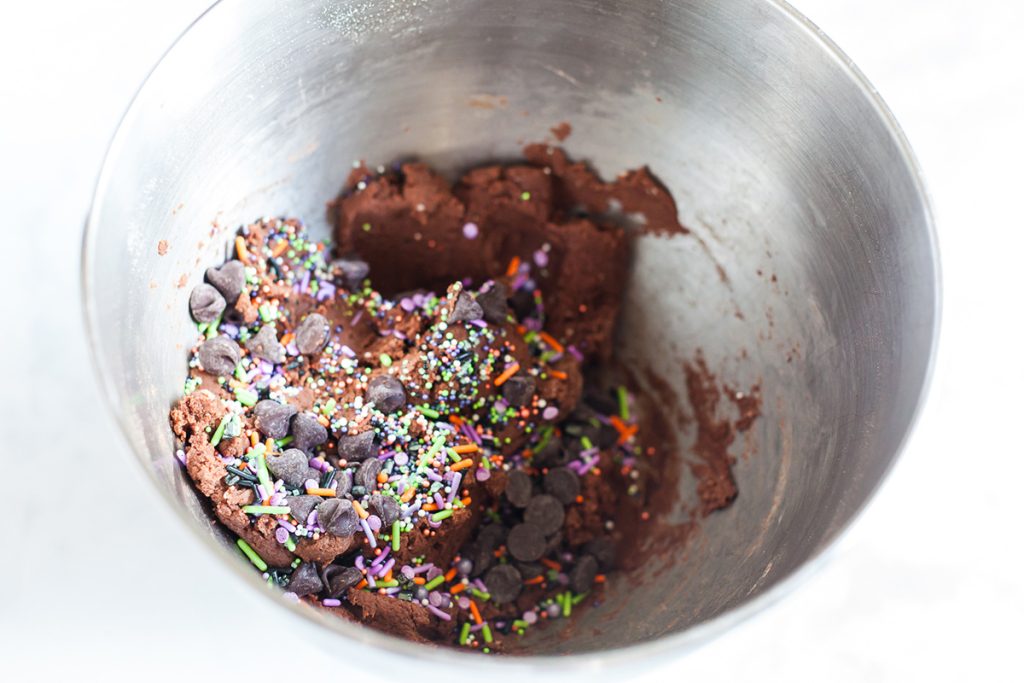 Cookie dough in a mixing bowl with chocolate chips and colorful Halloween sprinkles mixed in.