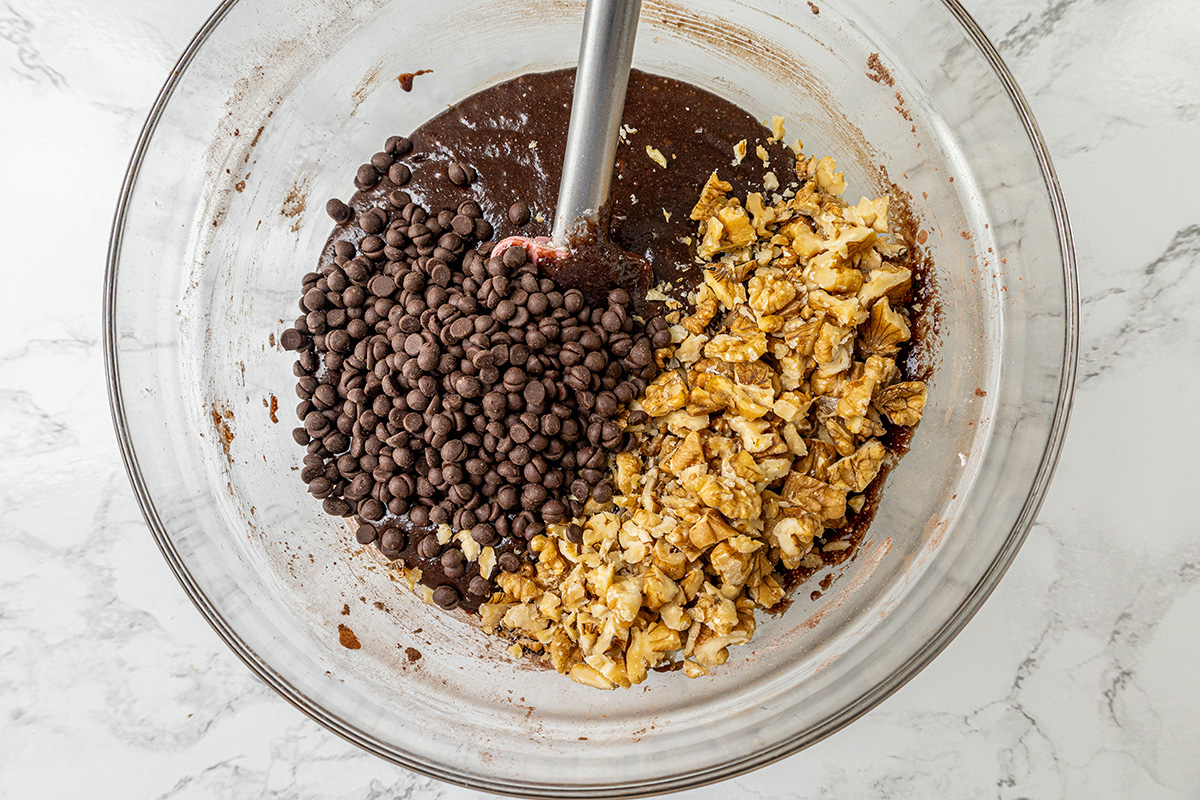 Chopped walnuts and chocolate chips poured into a bowl of chocolate brownie batter, ready to be mixed in.