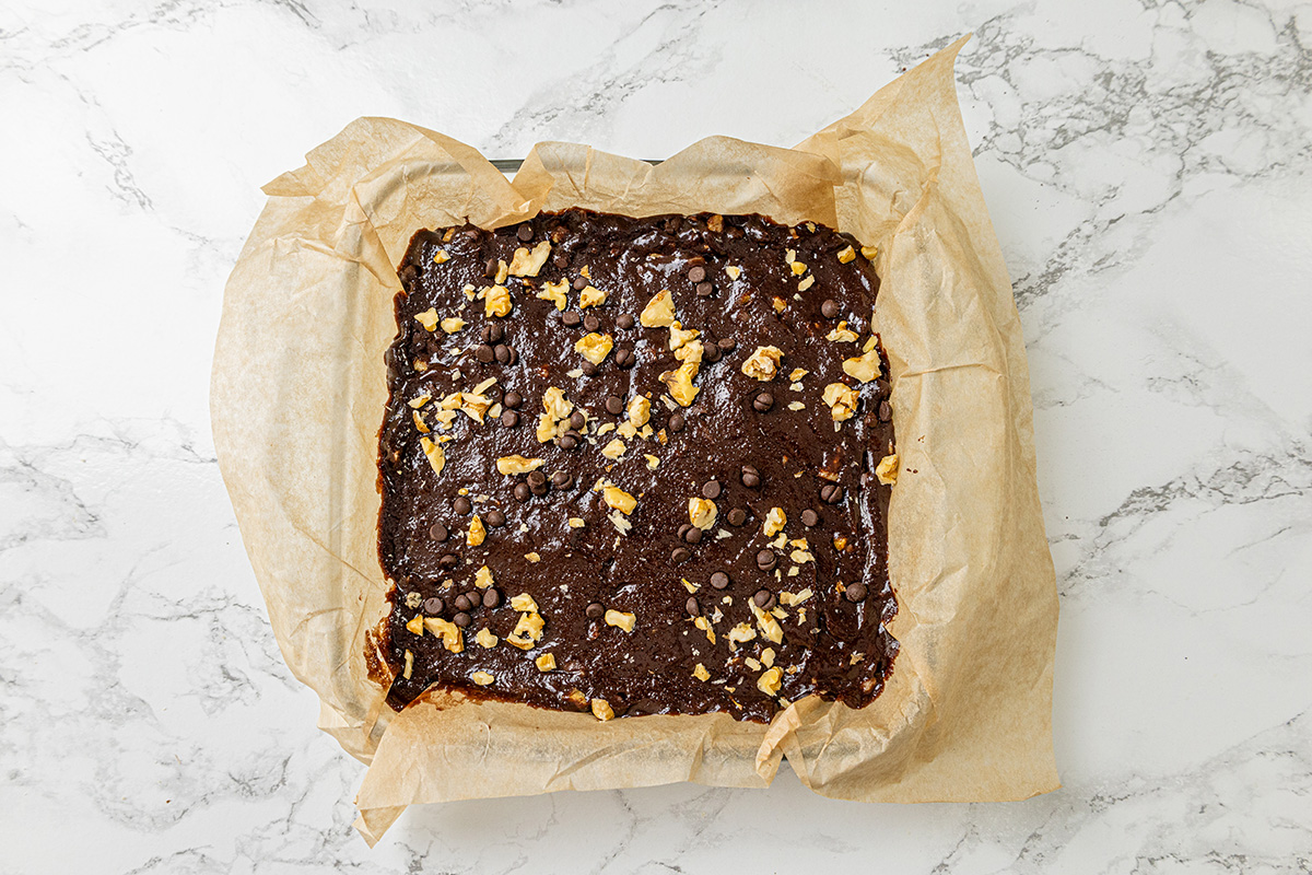 Chocolate brownie batter spread in a parchment-lined pan and topped with chopped walnuts and chocolate chips before baking.
