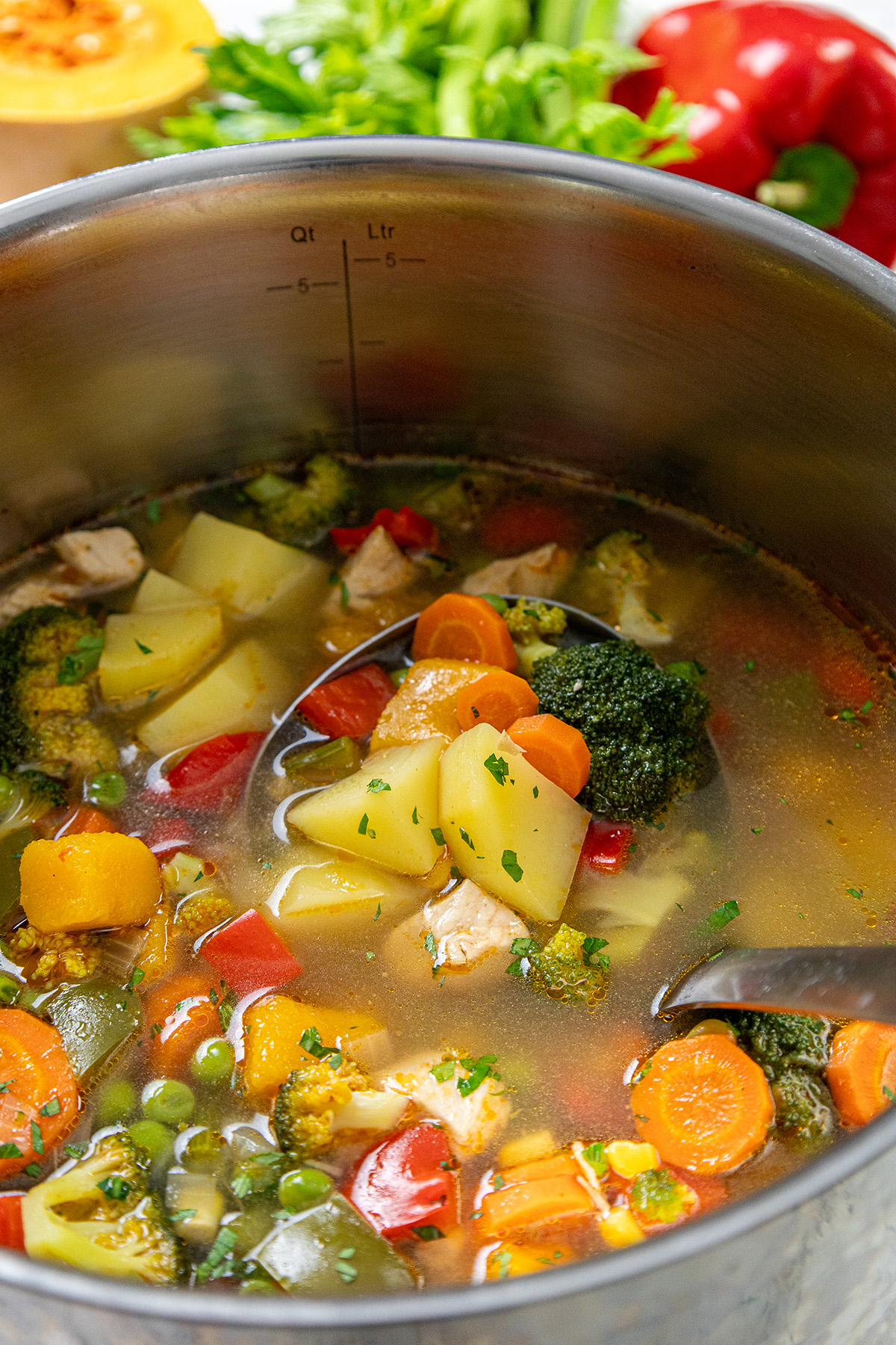 Ladle scooping chicken vegetable soup with potatoes, carrots, broccoli, and peppers from a large stainless steel pot.