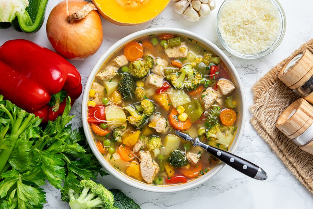 Bowl of chicken vegetable soup with potatoes surrounded by fresh vegetables like peppers, onion, celery, broccoli, and squash.
