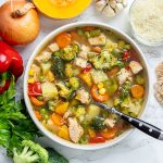 Bowl of chicken vegetable soup with potatoes surrounded by fresh vegetables like peppers, onion, celery, broccoli, and squash.