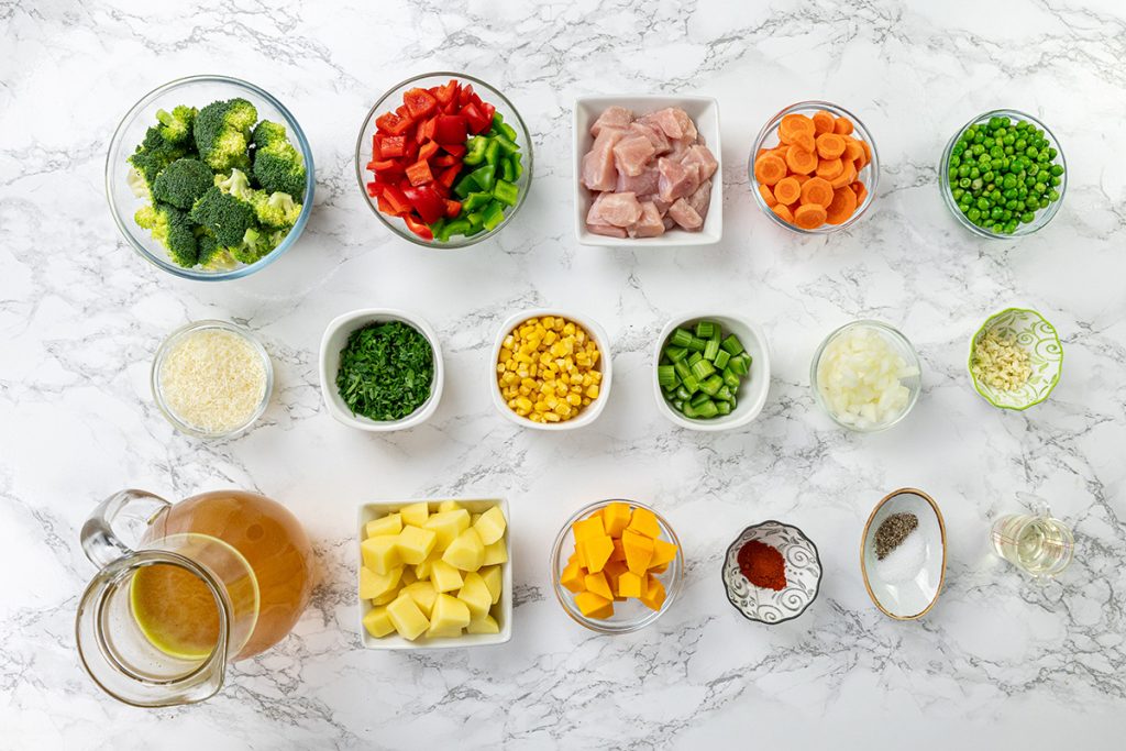 Ingredients prepped for chicken vegetable soup with potatoes, including broccoli, peppers, carrots, peas, corn, squash, potatoes, spices, broth, and cubed chicken.