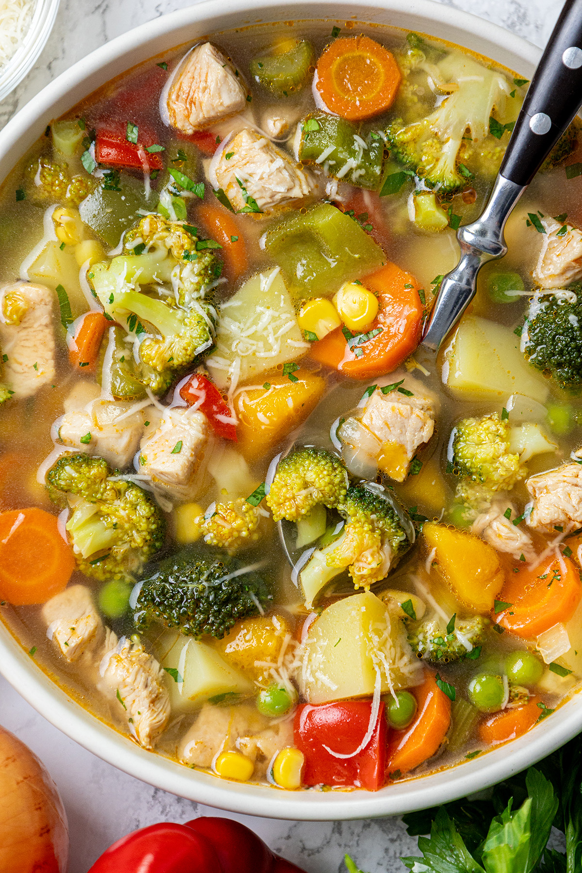 Close-up of homemade chicken vegetable soup with potatoes, carrots, broccoli, peppers, peas, and corn with shredded Parmesan on top.