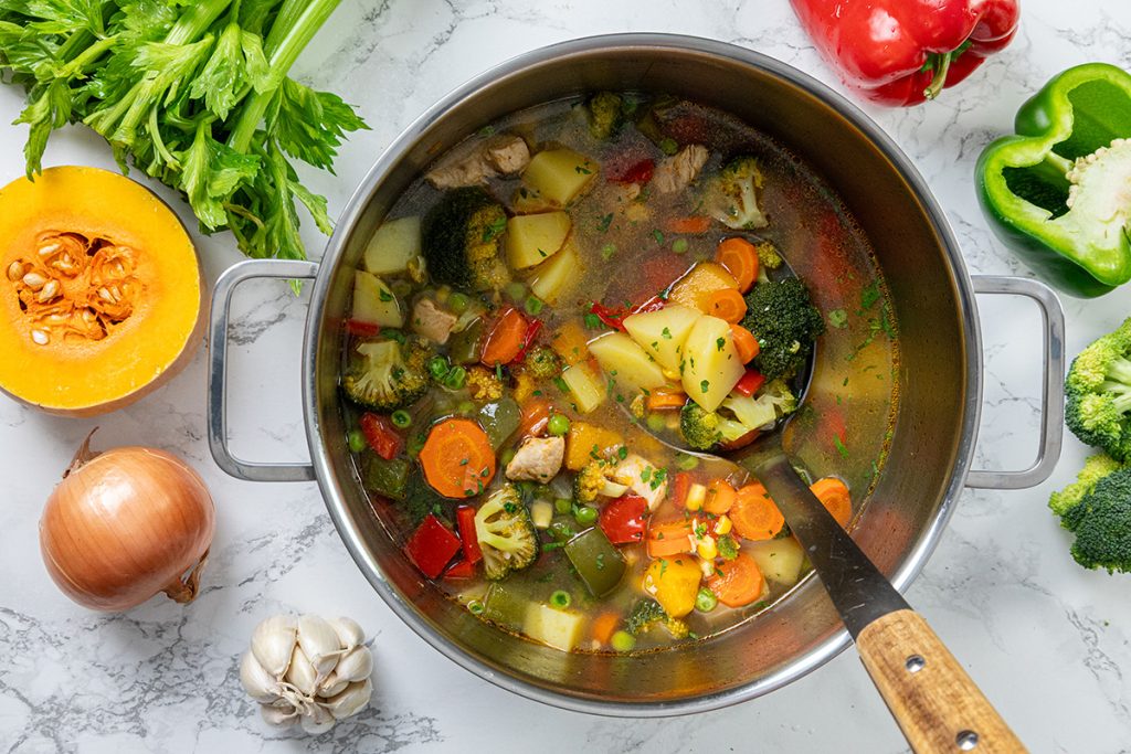 Homemade chicken vegetable soup with potatoes simmering in a pot, filled with carrots, broccoli, peppers, peas, and tender chicken.