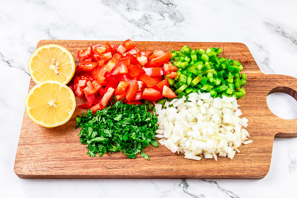 Chopped ingredients for Pebre on a wooden cutting board: tomatoes, green bell peppers, onions, cilantro, and halved lemons.