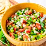 Yellow bowl of freshly made Pebre on a wooden board, surrounded by fresh cilantro and stacked tortillas.