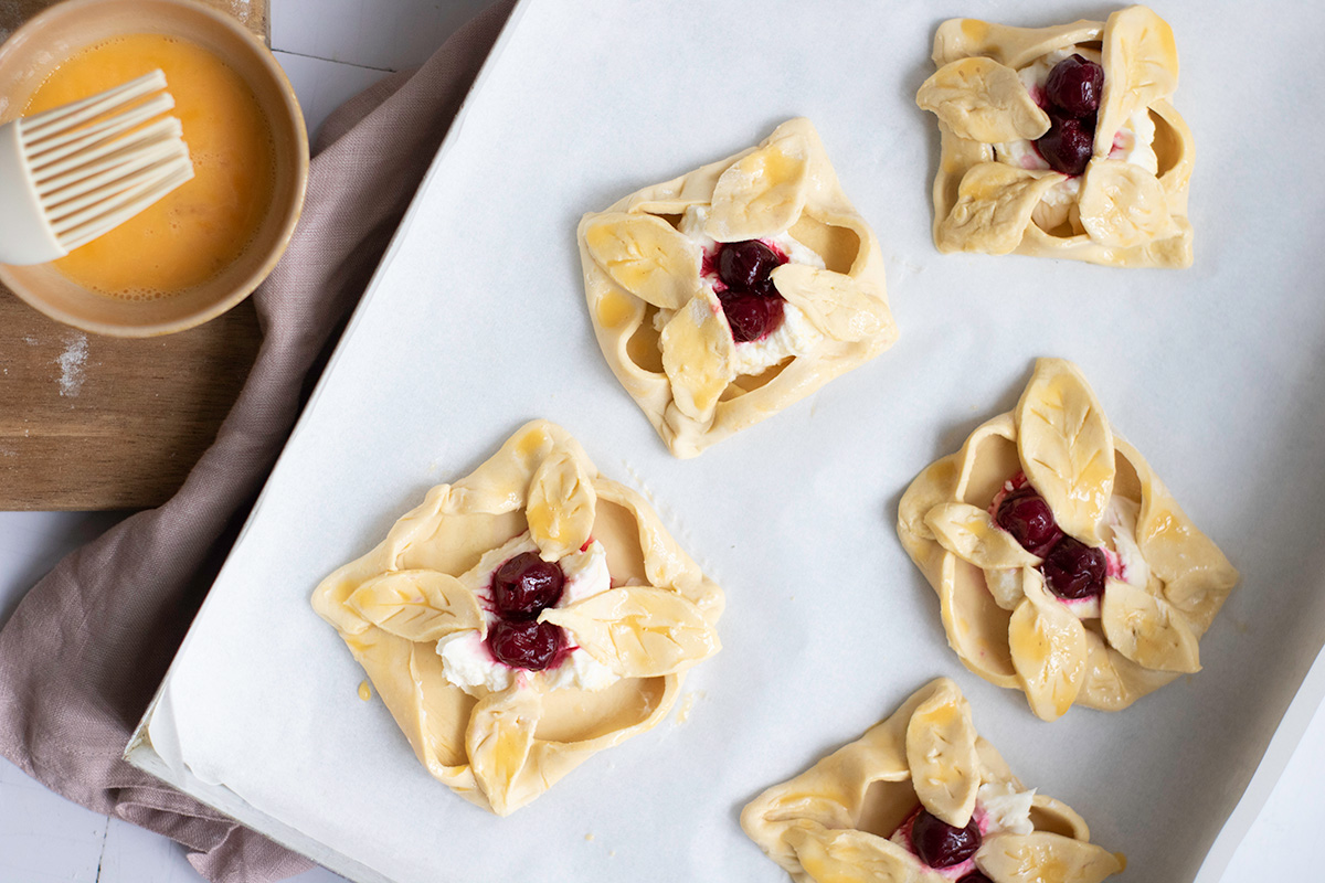 Showing cherry danish on a parchment paper lined baking sheet brushed with egg wash.
