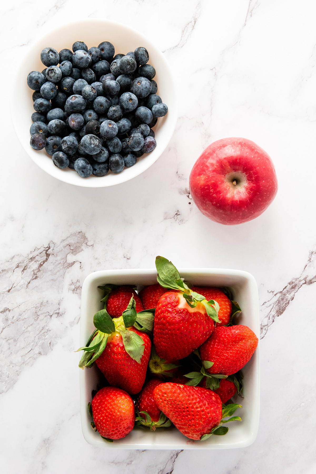 Fresh blueberries, strawberries, and a red apple arranged on a marble surface – perfect ingredients for a patriotic fruit salad.