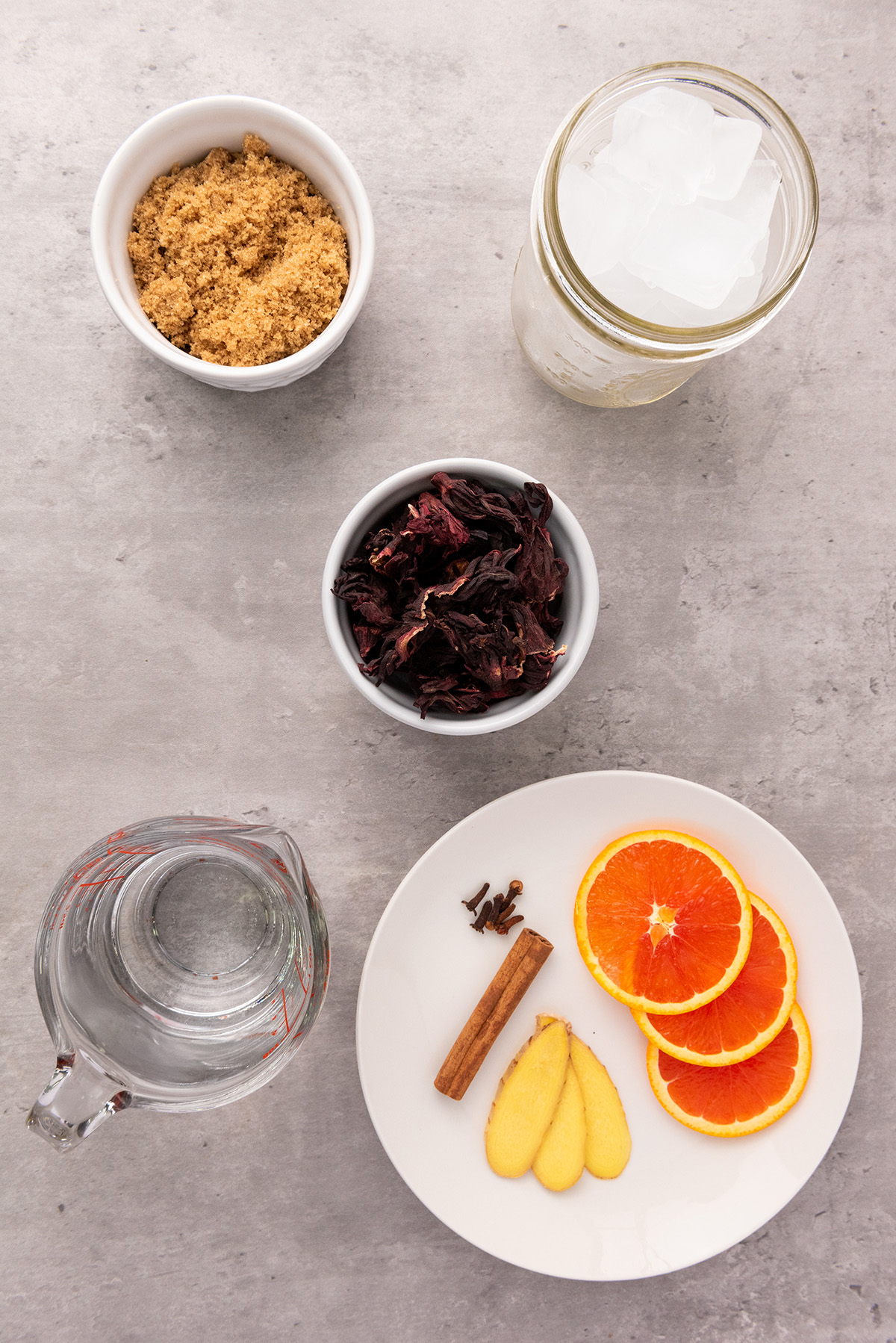 Ingredients for Agua de Jamaica laid out on a gray surface: dried hibiscus petals, brown sugar, ice cubes, water, orange slices, cinnamon stick, cloves, and fresh ginger.