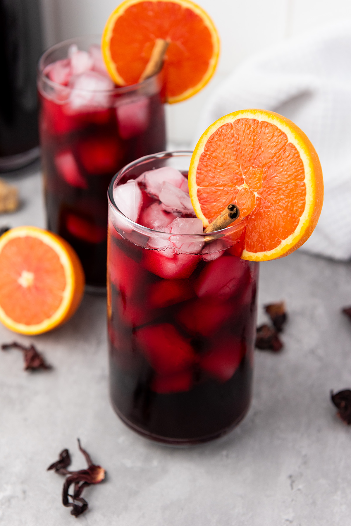Two tall glasses filled with iced hibiscus tea, each topped with an orange slice and cinnamon stick, with a bottle of tea in the background.
