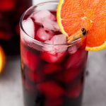 A glass of iced Agua de Jamaica garnished with a bright orange slice and cinnamon stick.