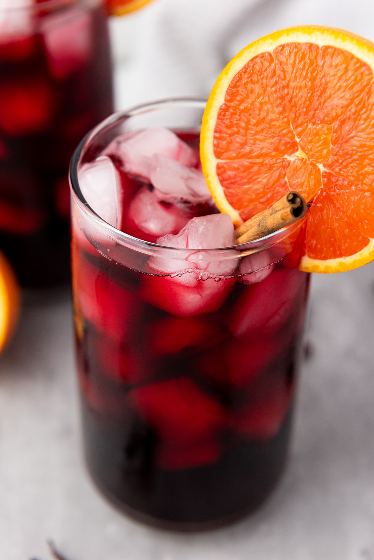 A glass of iced Agua de Jamaica garnished with a bright orange slice and cinnamon stick.