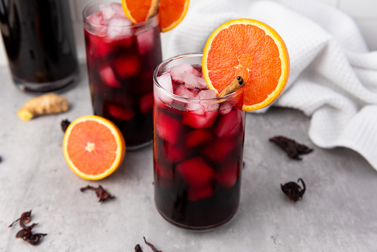 Close-up of two glasses of iced Agua de Jamaica on a gray surface, garnished with orange slices and surrounded by dried hibiscus petals.