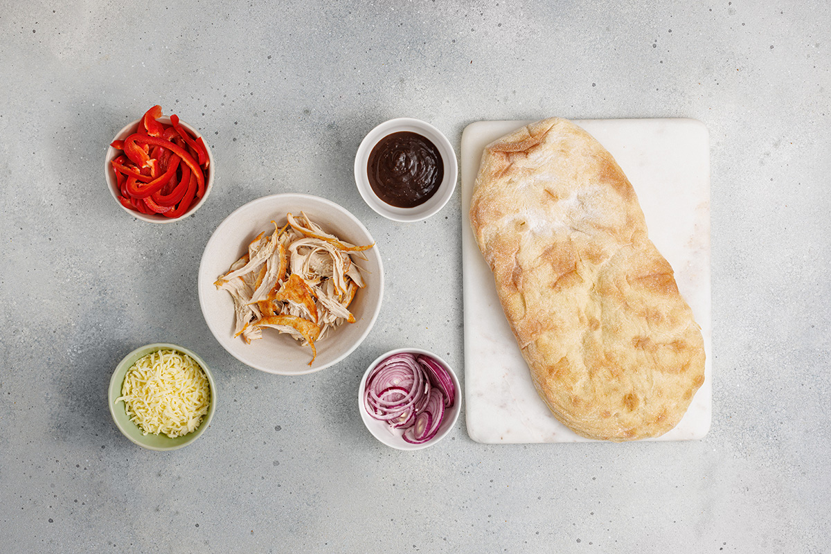 Ingredients for BBQ chicken flatbread arranged on a light surface, including shredded chicken, BBQ sauce, mozzarella cheese, sliced red onions, sliced red peppers, and flatbread.