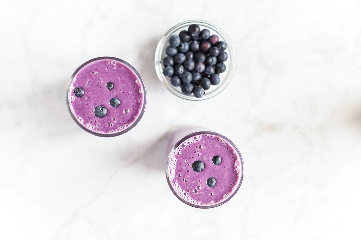 Overhead view of blueberry smoothies next to a bowl of fresh blueberries on a white surface.