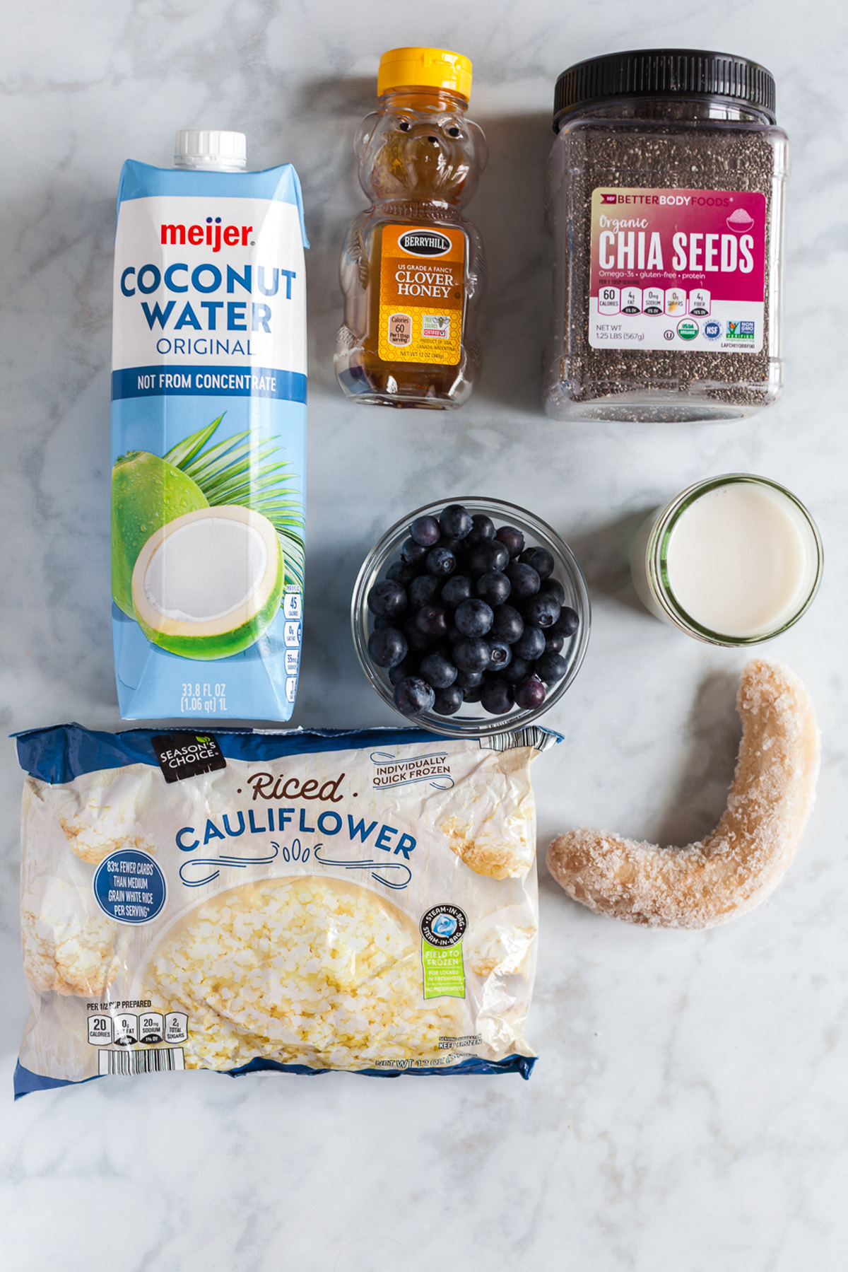 Flat lay of blueberry cauliflower smoothie ingredients including banana, chia seeds, coconut water, milk, honey, and frozen cauliflower.
