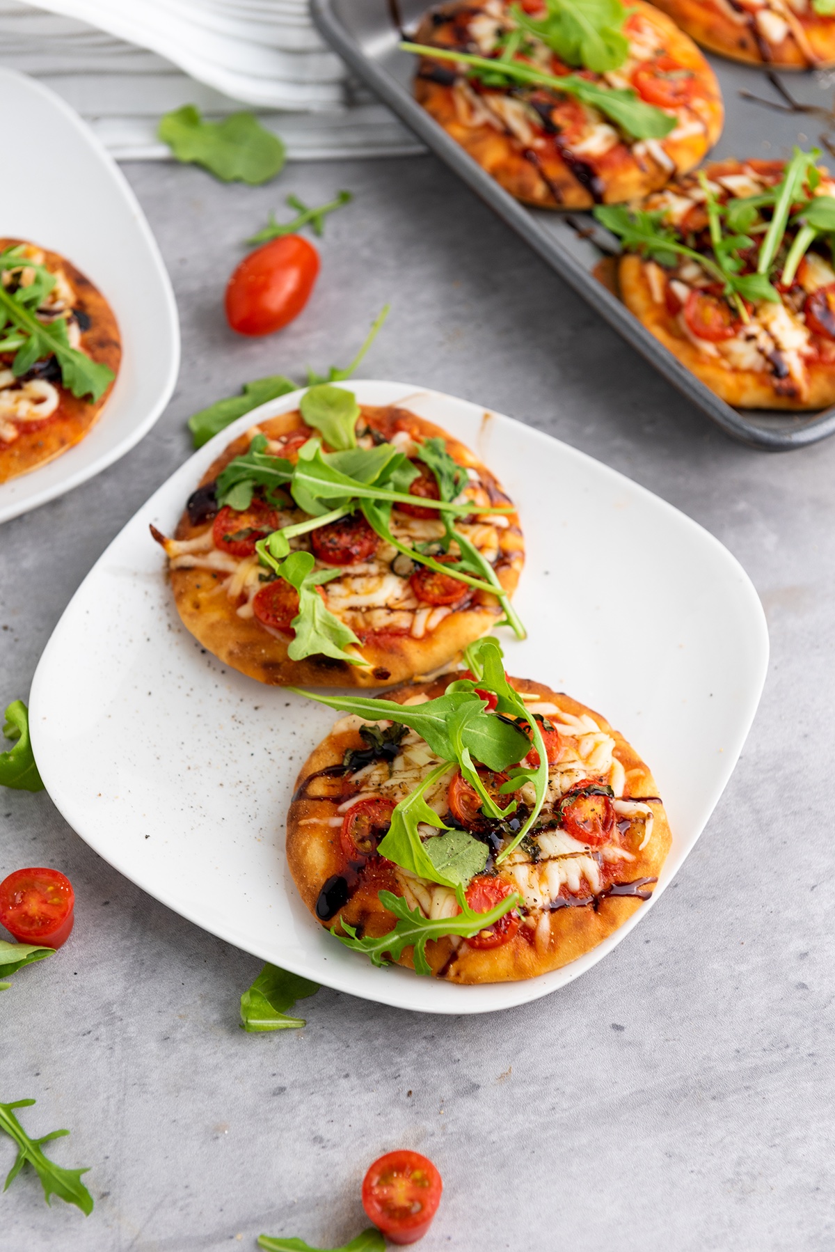 Mini Caprese naan pizzas topped with cheese, tomatoes, basil, and arugula arranged on a white plate and sheet pan for serving.