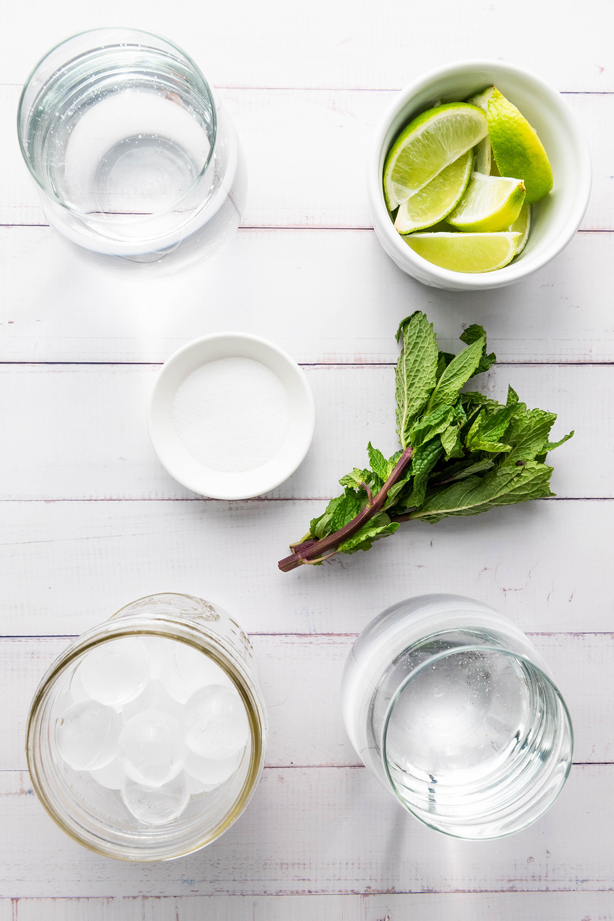 Flat lay of fauxjito ingredients: lime wedges, fresh mint, sparkling water, sugar, and ice on a white wood surface.