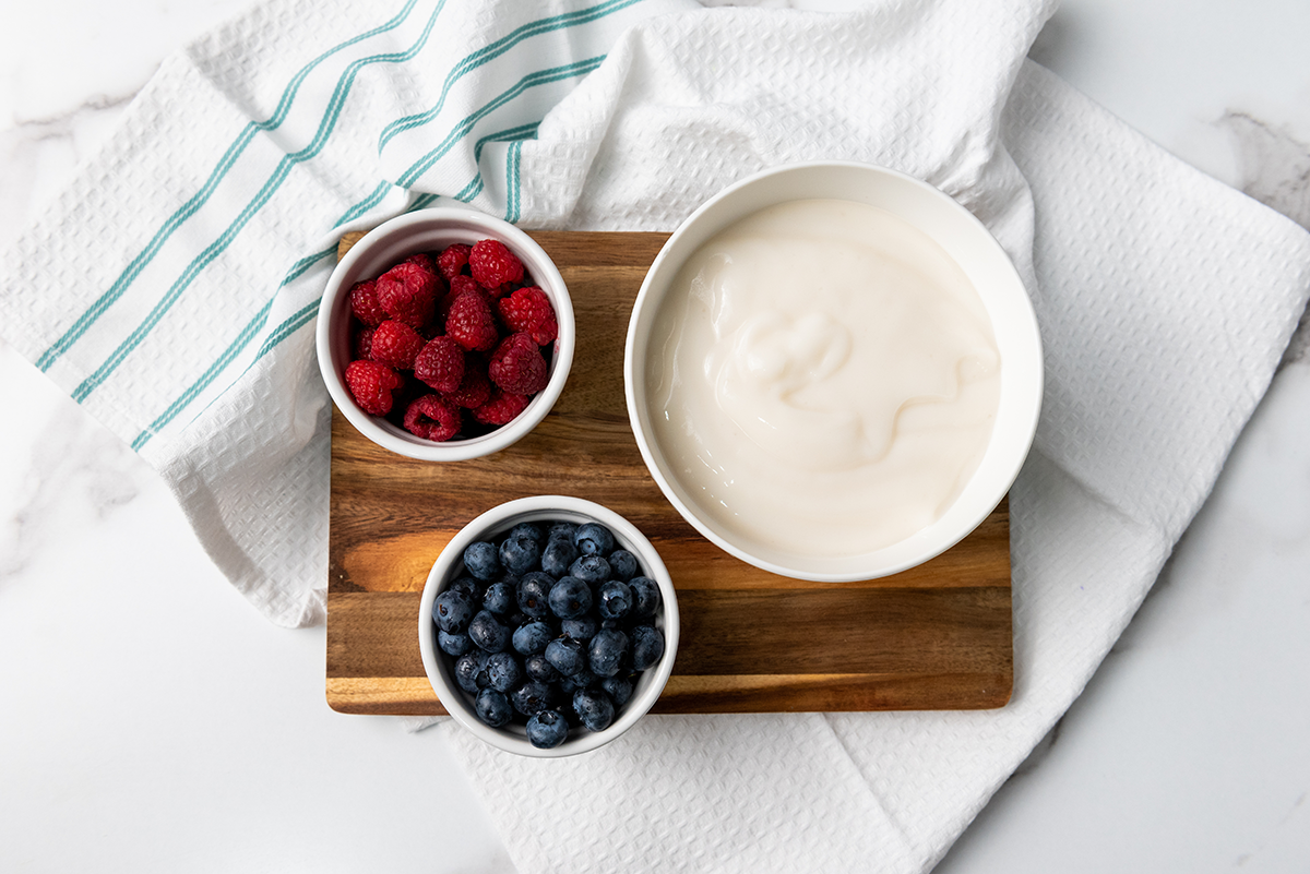 Three bowls on a wooden board with yogurt, fresh raspberries, and blueberries, ready to make frozen yogurt bites.
