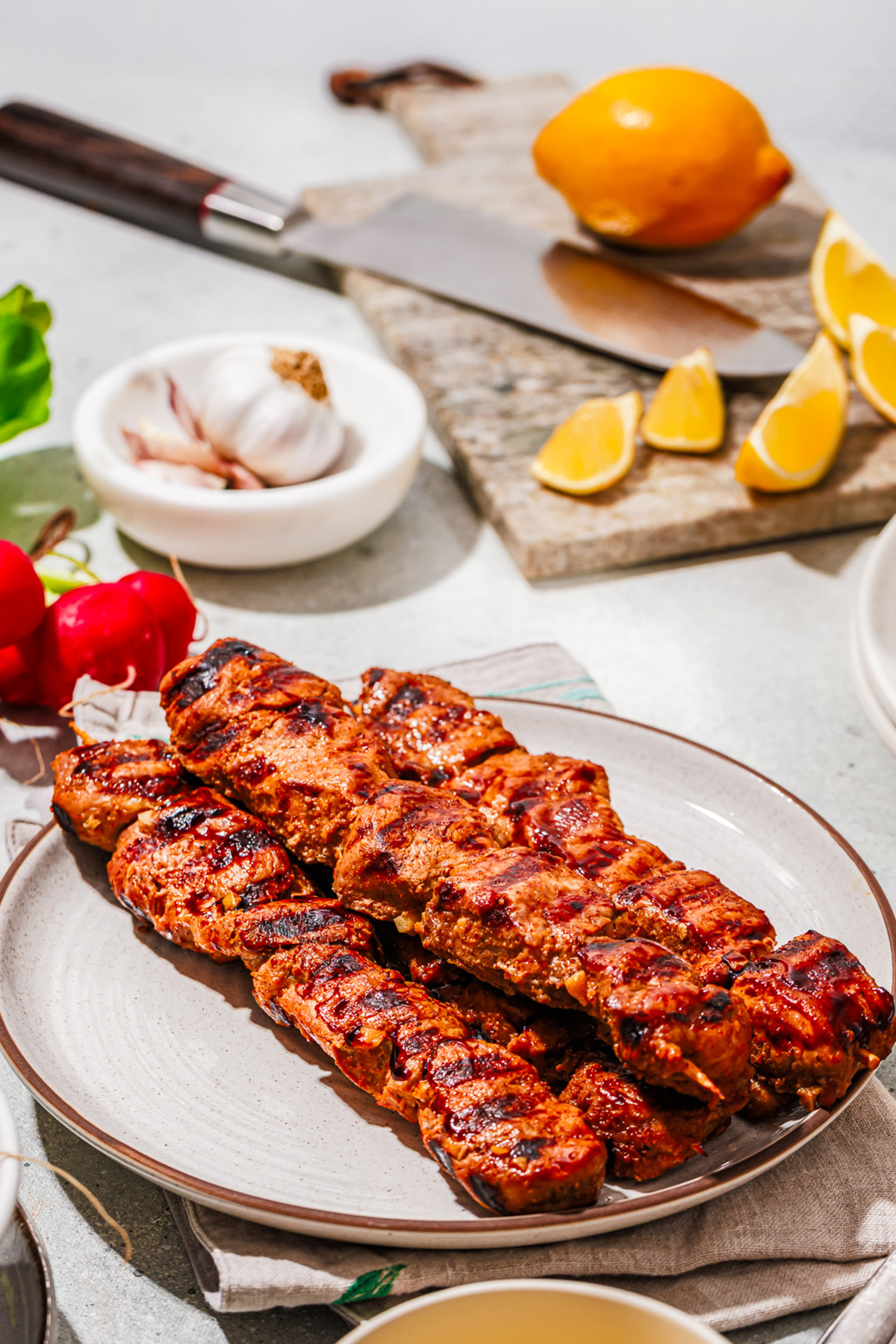 Top view of grilled pork skewers on a serving plate, surrounded by fresh summer ingredients like lemon and garlic.