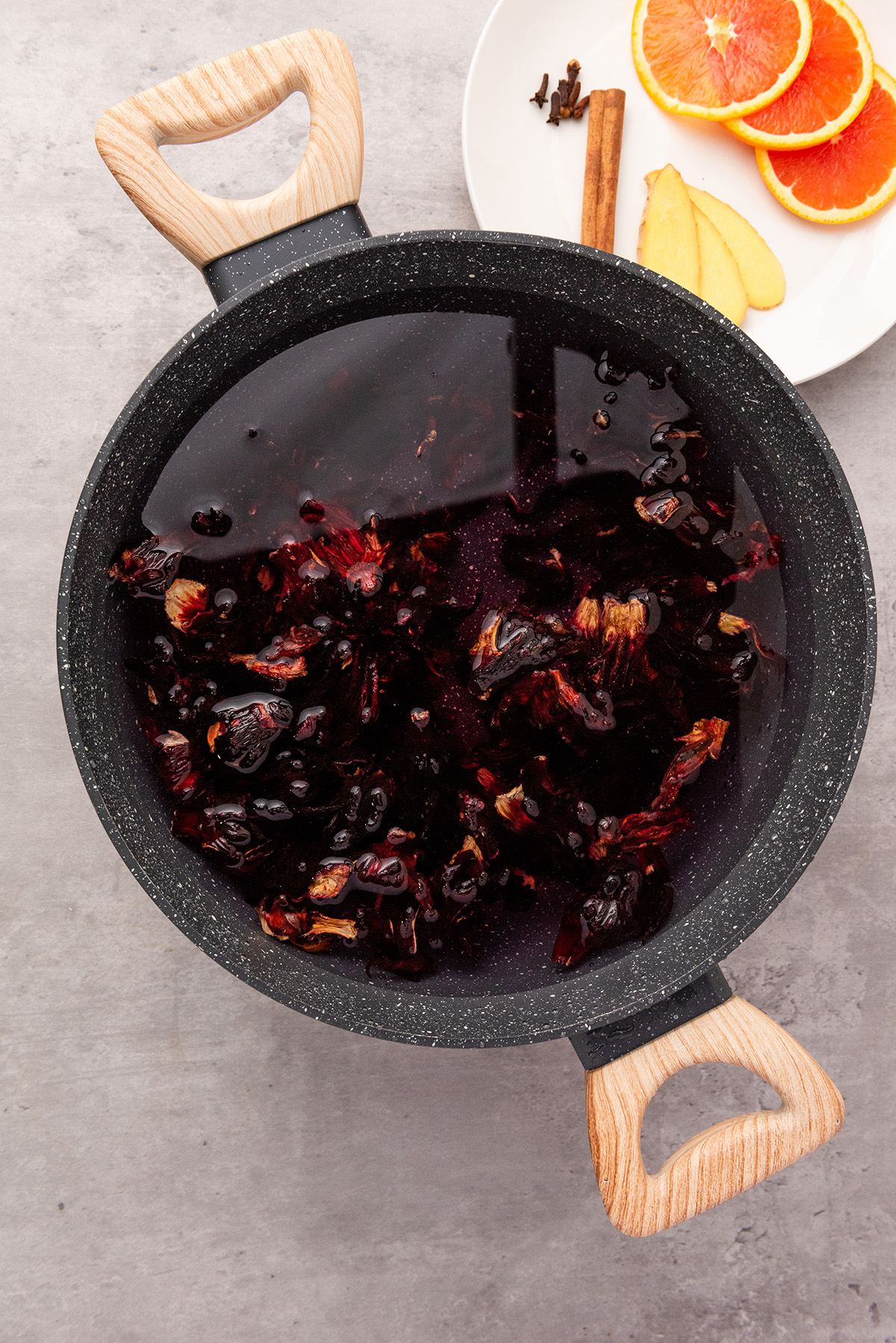A pot of hibiscus petals steeping in dark red liquid with a plate of fresh orange slices, cinnamon stick, cloves, and ginger nearby.