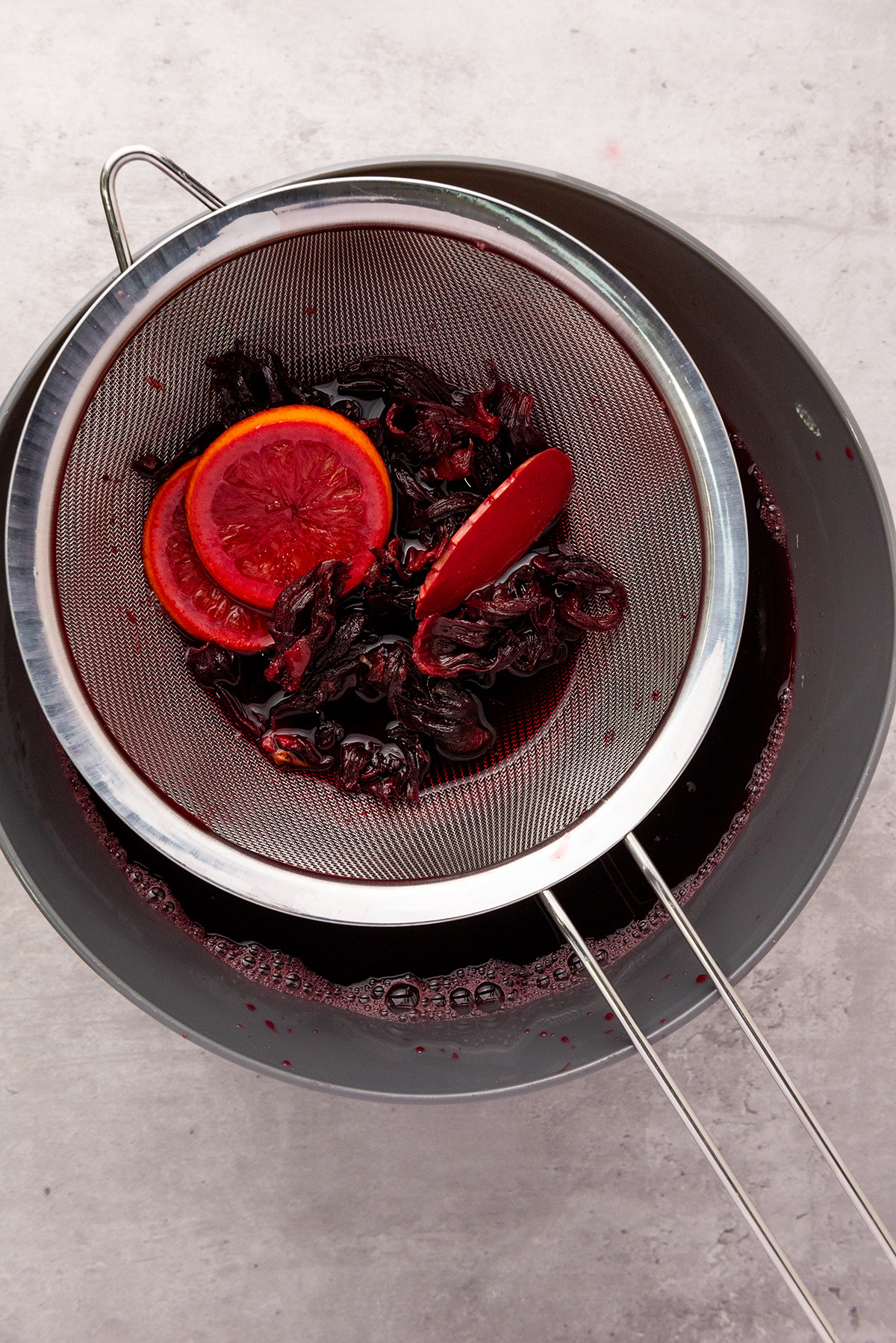 A fine mesh strainer resting over a pot, filled with steeped hibiscus petals and orange slices after brewing Agua de Jamaica.
