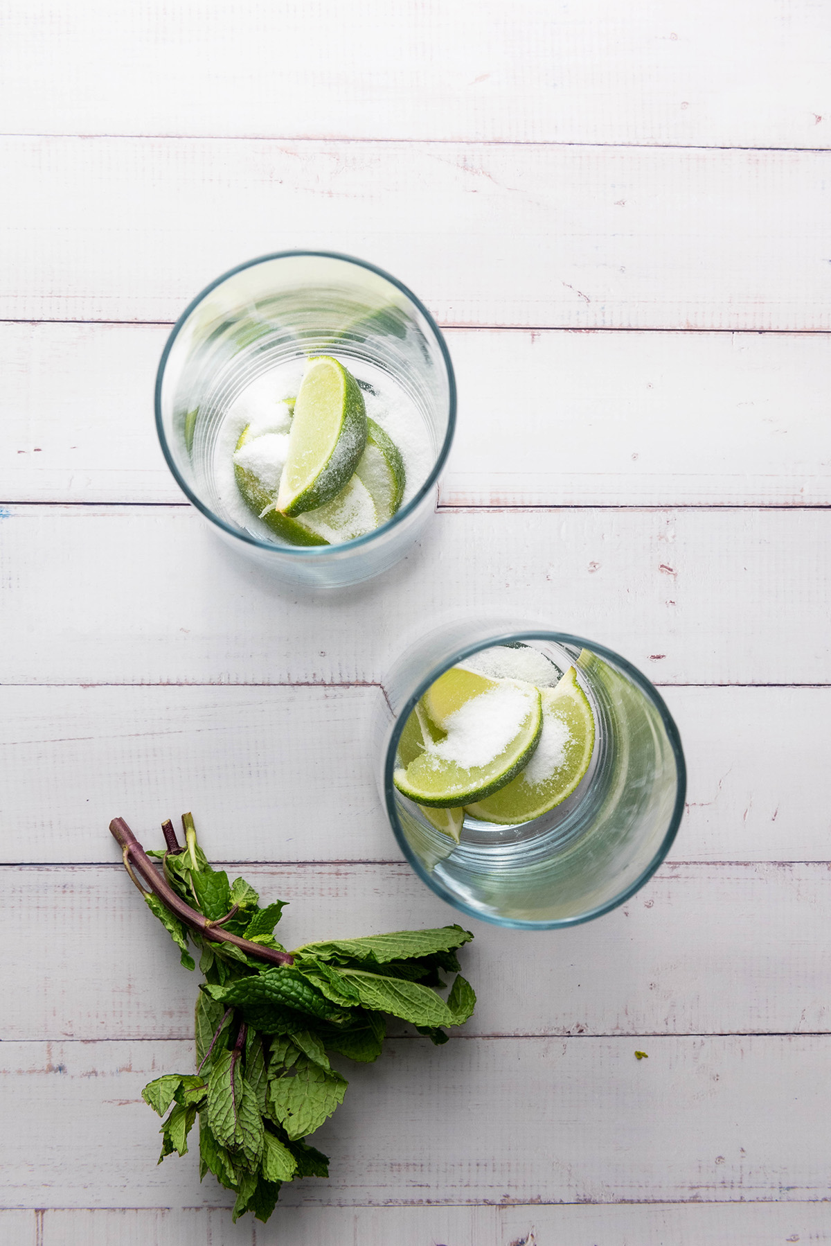 Two glasses with lime wedges and sugar at the bottom, fresh mint nearby, prepared for making fauxjitos