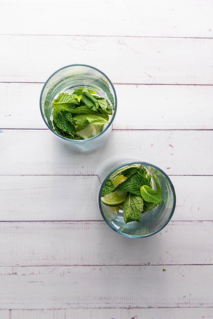Top-down view of two glasses filled with fresh mint and lime wedges on a white background, ready to be muddled.