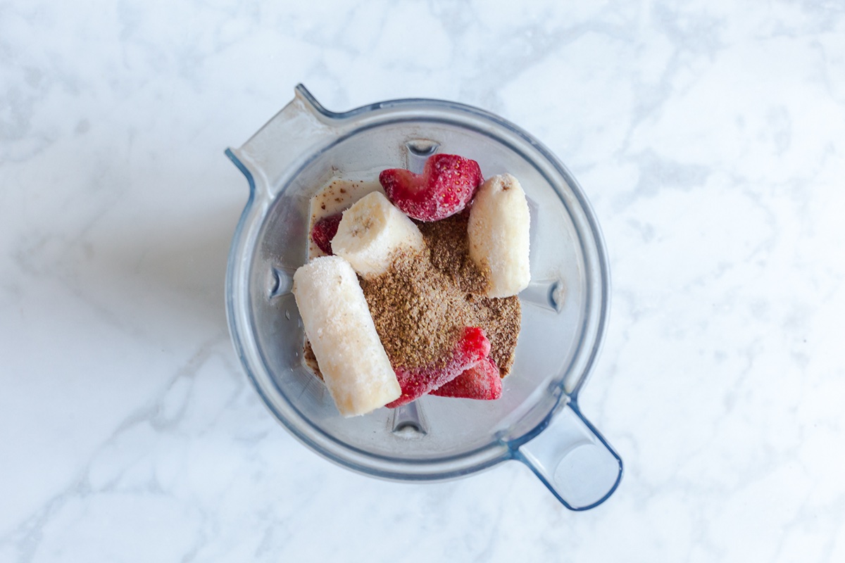  Overhead view of frozen banana, strawberries, flaxseed, and milk in a blender for strawberry banana flaxseed smoothie.
