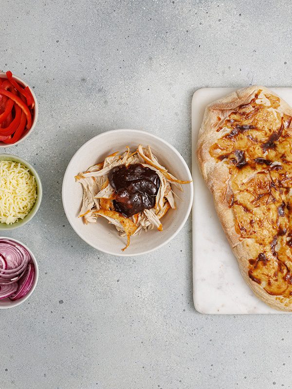 Shredded chicken being mixed with BBQ sauce in a bowl, with one flatbread brushed with sauce beside it.