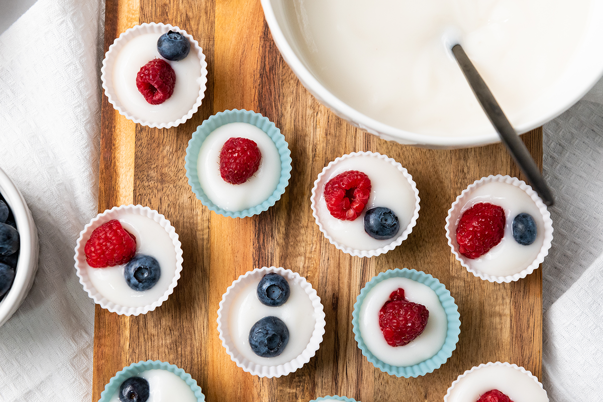Mini yogurt cups topped with fresh raspberries and blueberries, arranged on a wooden board before freezing.