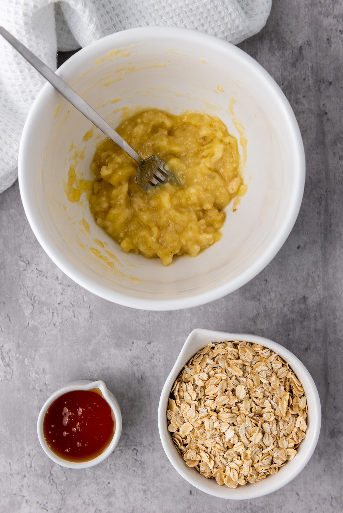 Mashed ripe banana in a white mixing bowl with a fork, ready to be combined with oats and honey.