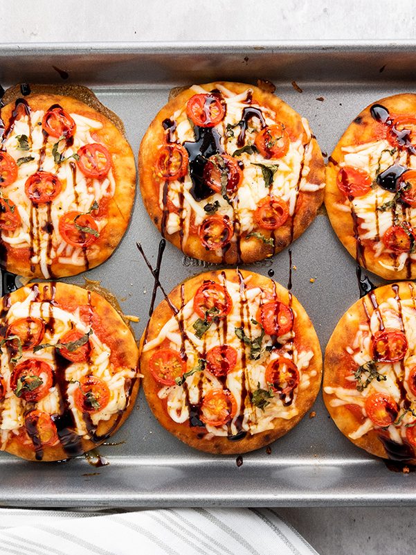 Baked mini Caprese pizzas fresh out of the oven on a sheet pan, drizzled with balsamic glaze and golden around the edges.