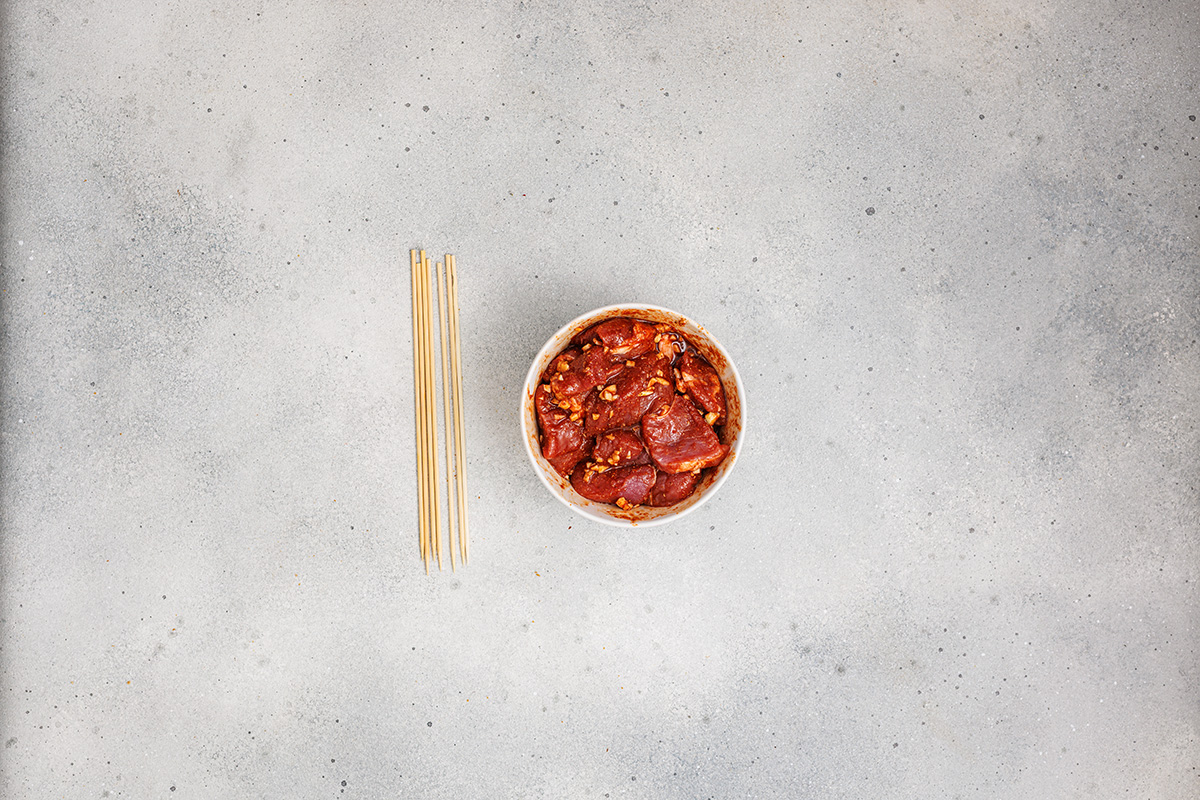 Bowl of marinated pork tenderloin chunks beside a bundle of wooden skewers on a light background.