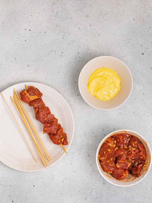 Single skewer of marinated pork on a plate, with more marinade and aioli nearby, ready for grilling.