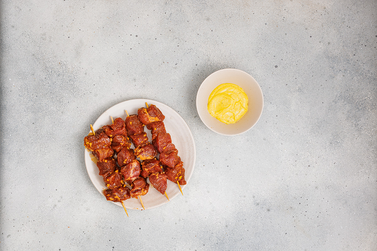 Top-down view of multiple pork skewers on a plate with a bowl of aioli, prepped for the grill.