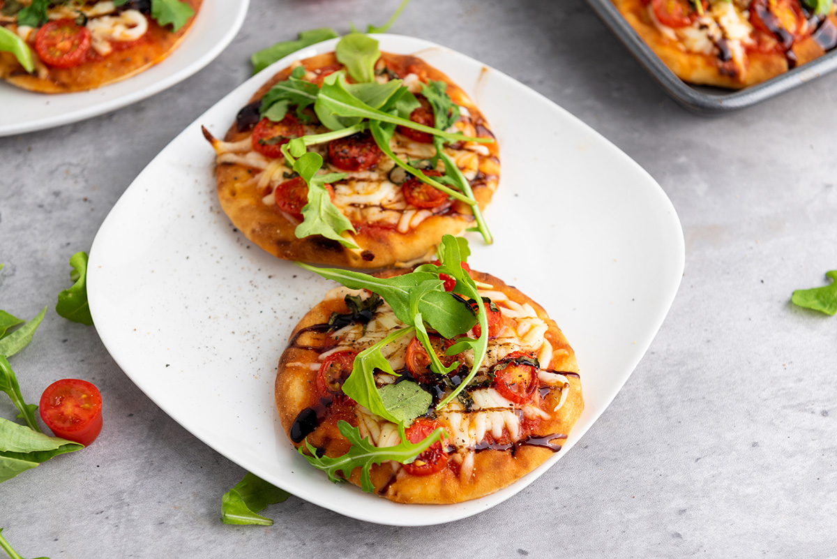 Two mini Caprese naan pizzas served on a plate with scattered arugula and cherry tomato halves nearby, perfect for a party appetizer.