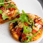 Close-up of two mini Caprese naan pizzas topped with melted mozzarella, cherry tomatoes, fresh arugula, and a balsamic glaze drizzle on a white plate.