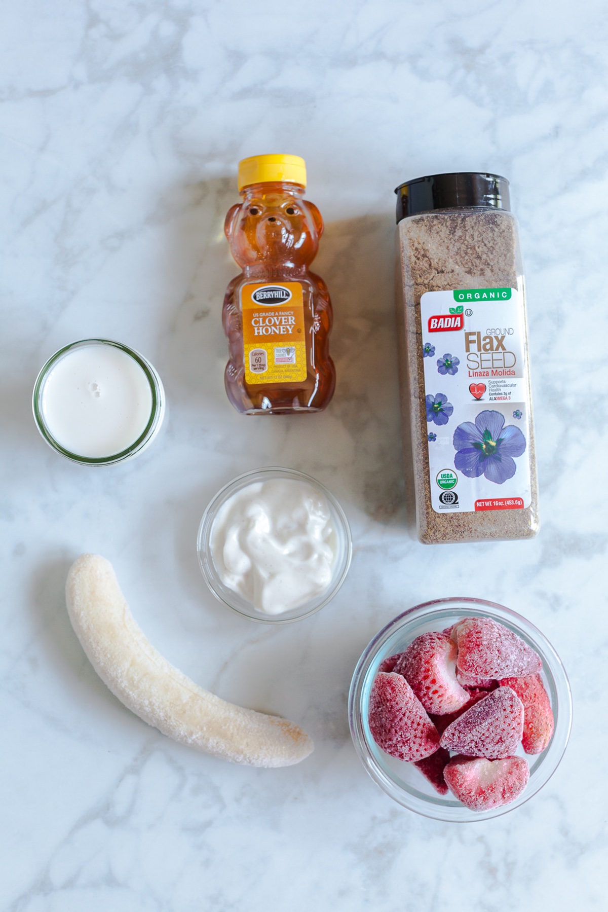 Flat lay of ingredients for strawberry banana flaxseed smoothie including frozen banana, strawberries, ground flaxseed, yogurt, honey, and milk.