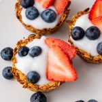 Overhead shot of three yogurt granola cups topped with fruit, surrounded by loose blueberries and strawberries.