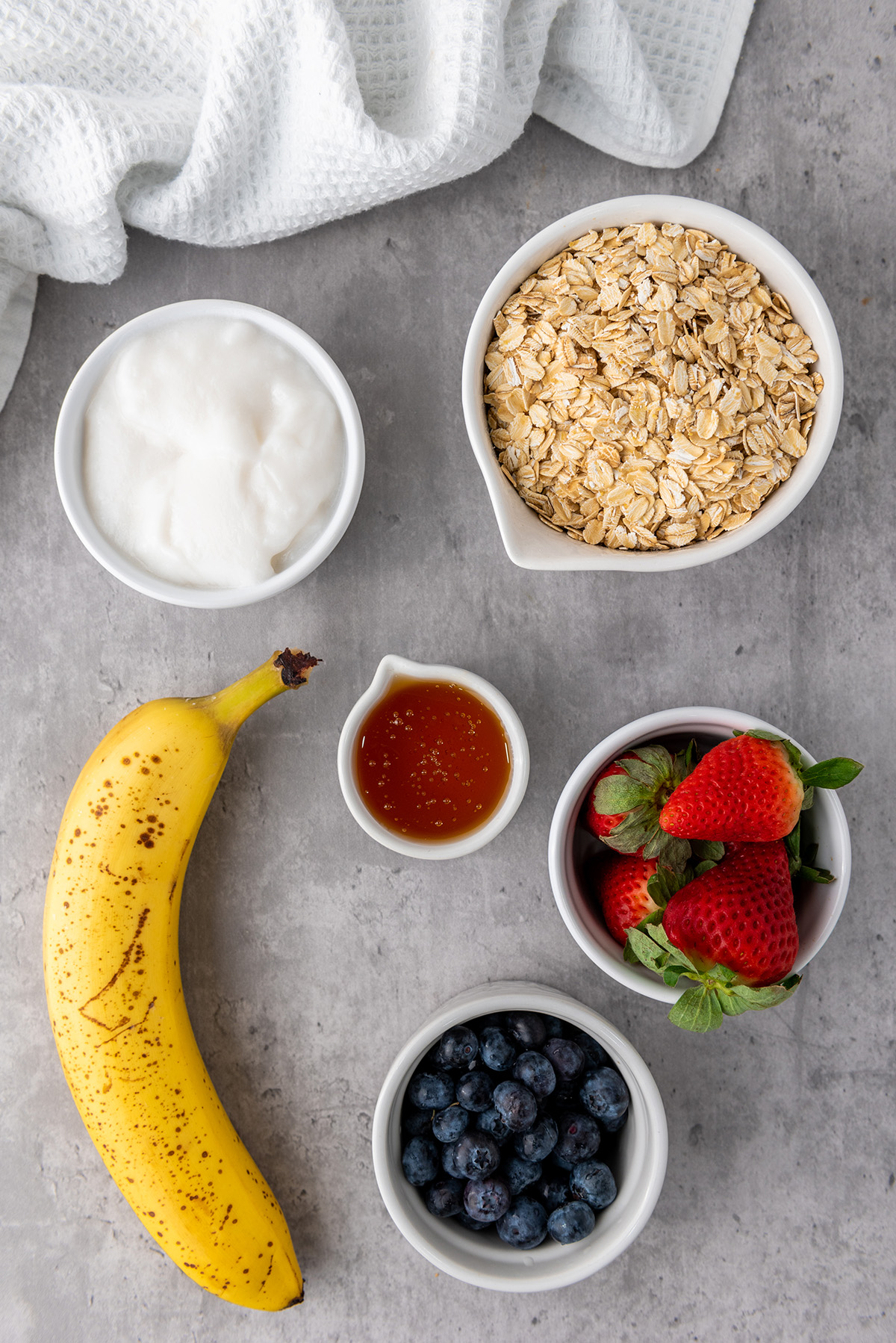 Overhead view of fresh ingredients for yogurt granola cups including a banana, oats, yogurt, honey, strawberries, and blueberries on a gray surface.