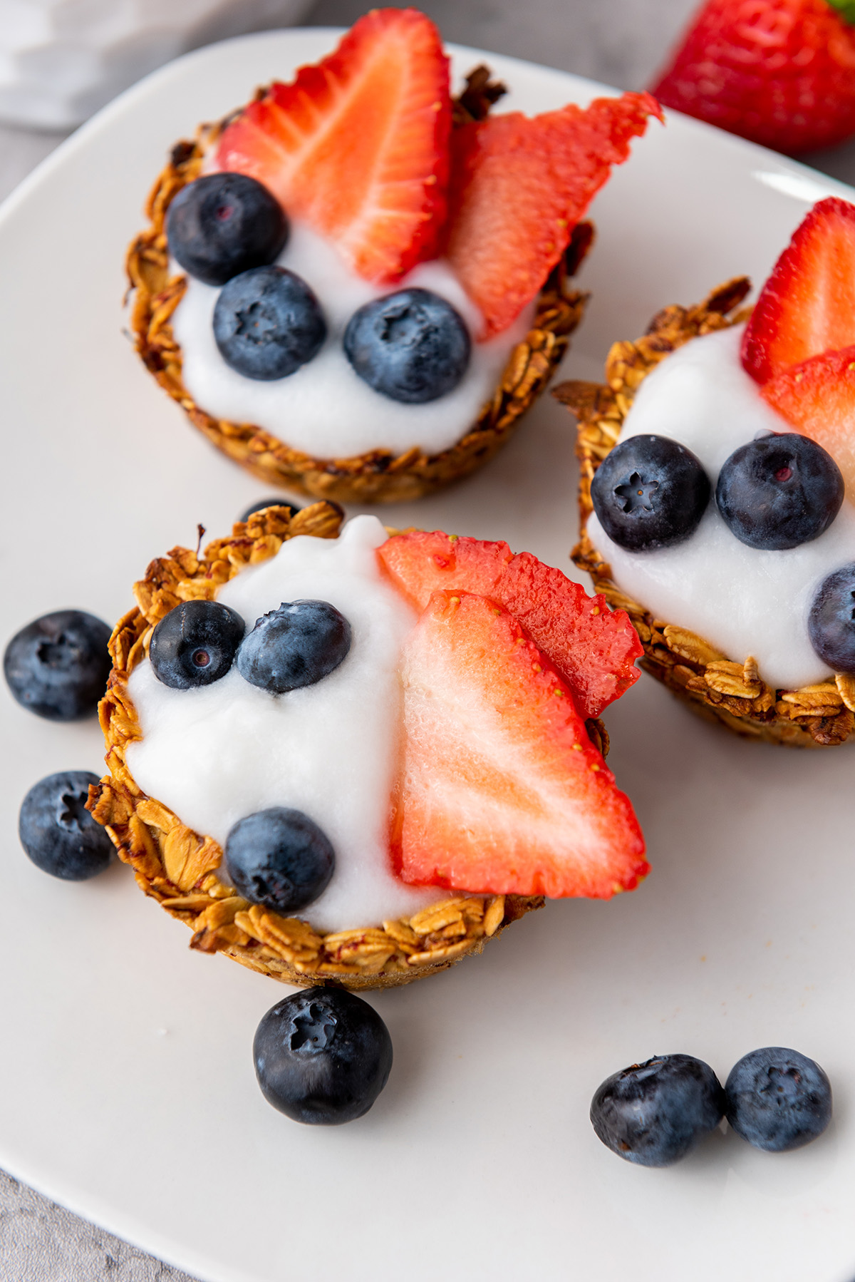 Overhead shot of three yogurt granola cups topped with fruit, surrounded by loose blueberries and strawberries.