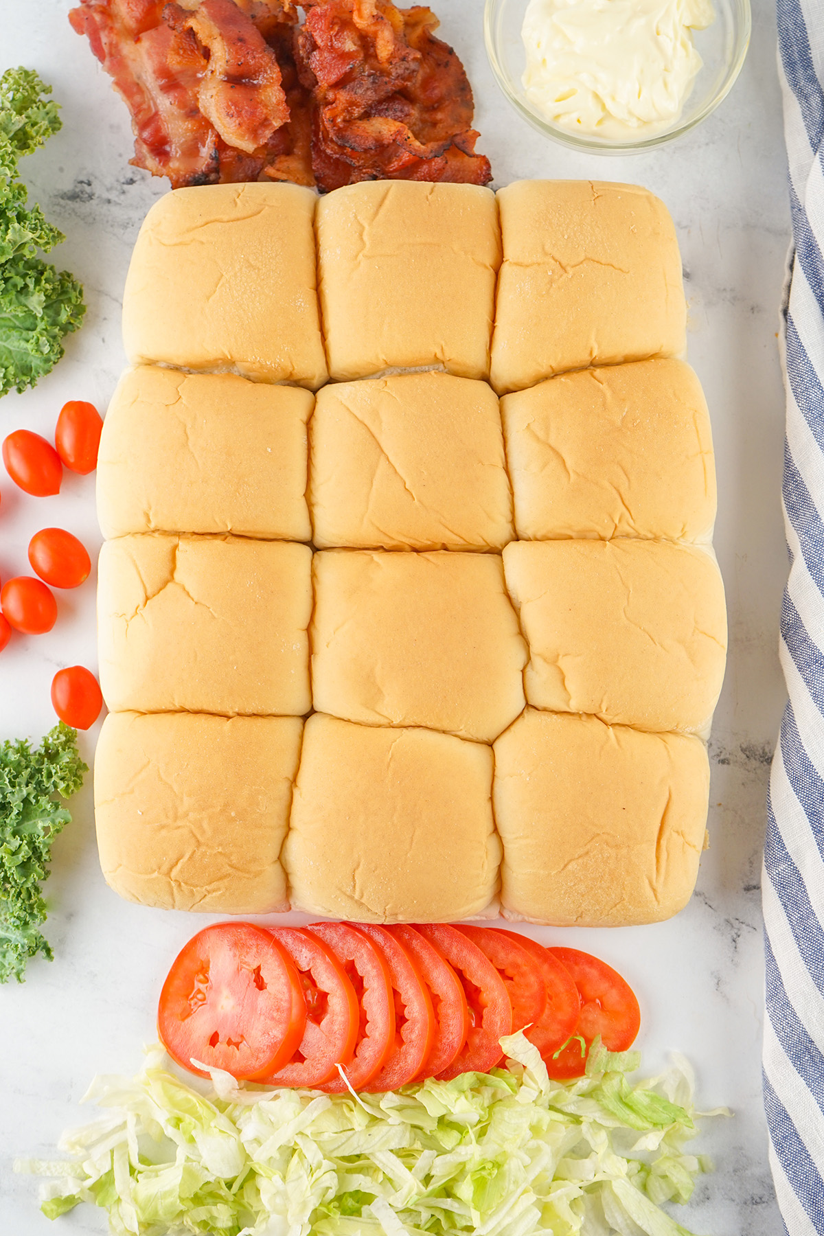 Full sheet of soft Hawaiian rolls before slicing, surrounded by sandwich ingredients, bacon, tomato, lettuce, and mayo.